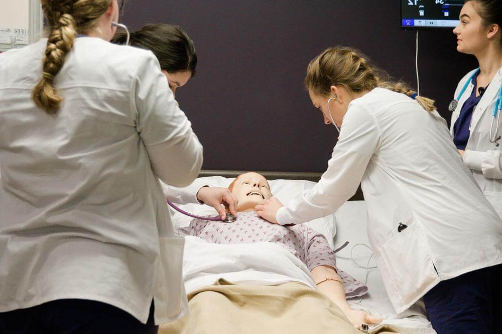 Nursing students watch as female nursing student practices taking vital signs from mannequin laying in hospital bed