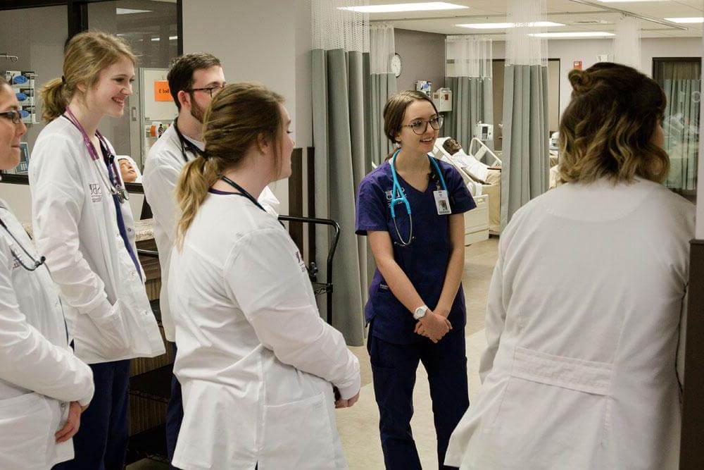 Nursing students in scrubs stand in simulation lab listening to professor