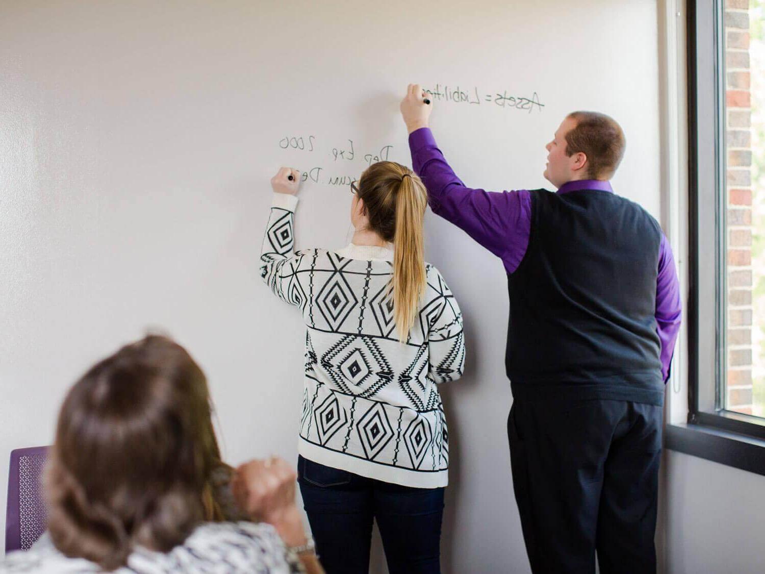 two students practice accounting problems on whiteboard as professor looks on