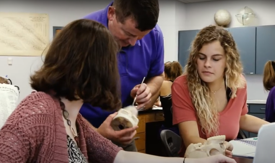 female students look on as biology professor points to model to explain experiment
