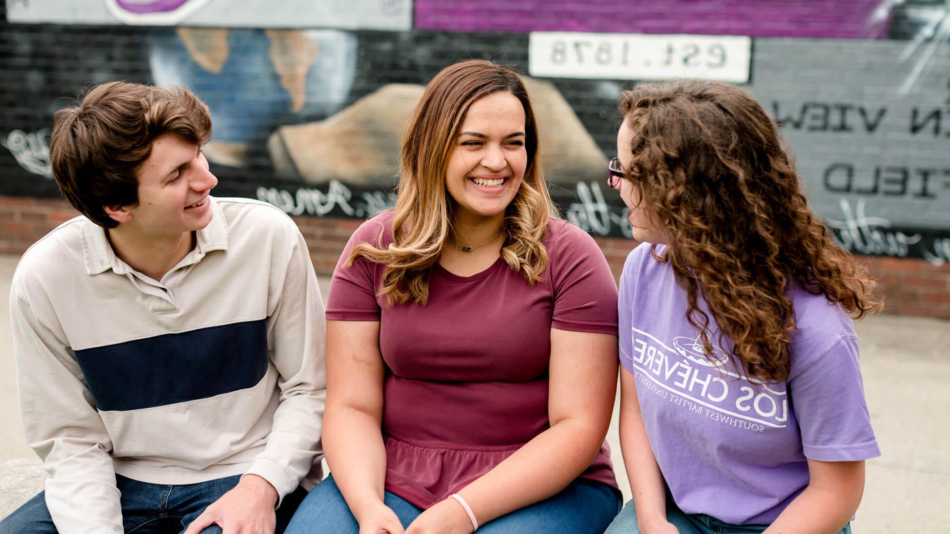 three students smiling while sitting in front of mural on brick wall