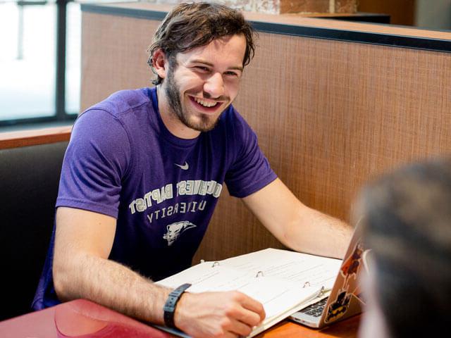 Male student smiles while sitting in booth with book open on table in front of him